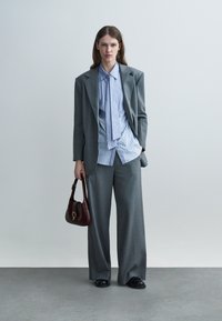 Femme debout portant un costume gris à jambes larges, une chemise rayée bleue avec un détail de cravate, des chaussures noires, tenant un sac à main marron foncé sur un fond uni.