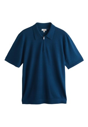 Mand med gråt skæg iført en marineblå poloshirt med korte ærmer, hvide plisserede bukser og mørkebrune sko, der står indendørs.