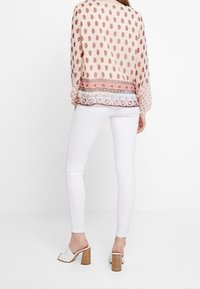 Blusa a maniche lunghe, a fantasia in pesca chiaro con motivi ovali marroni e rifiniture in pizzo, abbinata a jeans skinny bianchi e mule bianchi con tacco.