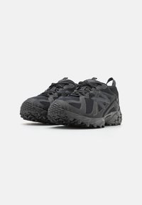 Schwarze Sportschuhe mit Mesh-Obermaterial, Gummisohle, strukturiertem Profil und verstärktem Zehenbereich. Mit Schnürsenkeln und Zuglasche an der Ferse.