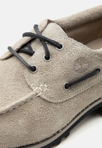 Bézs színű velúr fűzős cipő fekete fűzőkkel, részletes varrással és fa logóval az füzőlyukaknál.