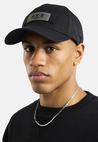 Czarny czapka bejsbolowa z teksturowanym materiałem i prostokątną naszywką z napisem "A.V.T" w odcieniu szarości. Model nosi czarny sweter i srebrny łańcuszek.