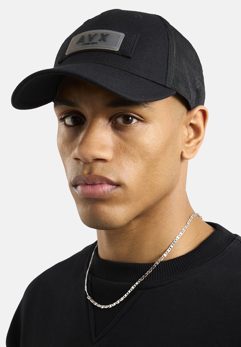 Czarny czapka bejsbolowa z teksturowanym materiałem i prostokątną naszywką z napisem "A.V.T" w odcieniu szarości. Model nosi czarny sweter i srebrny łańcuszek.