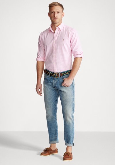 Camisa de cuadros rosa de botones, con las mangas remangadas; combinada con jeans azules desgastados, zapatos de barco de cuero marrón y un cinturón multicolor.