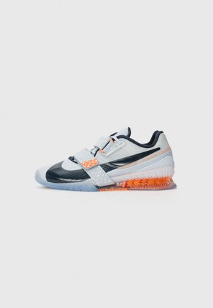 Chaussure de sport blanche, noire et orange avec deux sangles, semelle texturée et amorti orange visible au niveau du talon sur un fond gris.