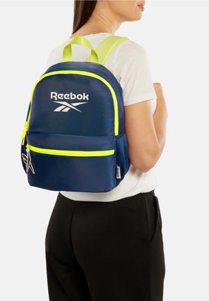 Mujer con mochila Reebok azul marino con cremalleras y correas amarillas neón, vestida con camisa blanca y pantalones negros, mirando hacia la derecha.