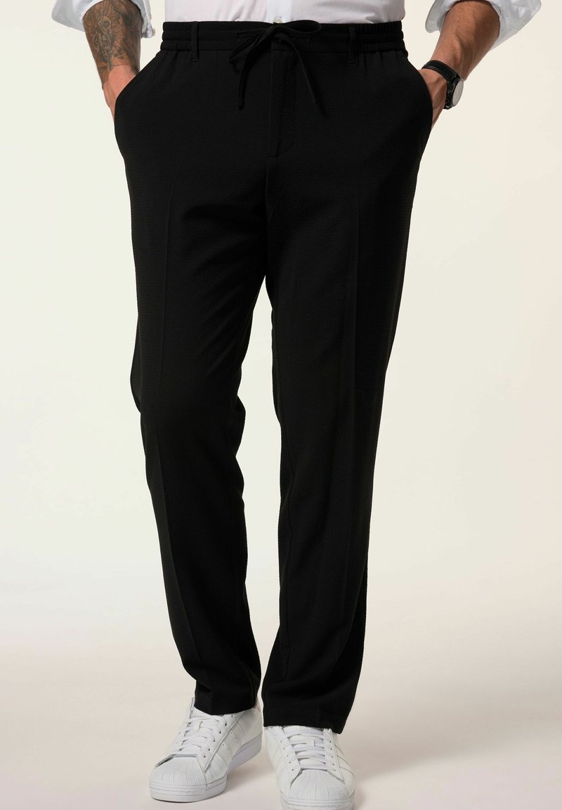 Mustat housut, jotka on valmistettu teksturoidusta kankaasta, varustettuna joustavalla vyötärönauhalla ja nyörillä, sivutaskuilla ja slim fit -mittasuhteilla, yhdistettynä valkoisiin lenkkareihin.