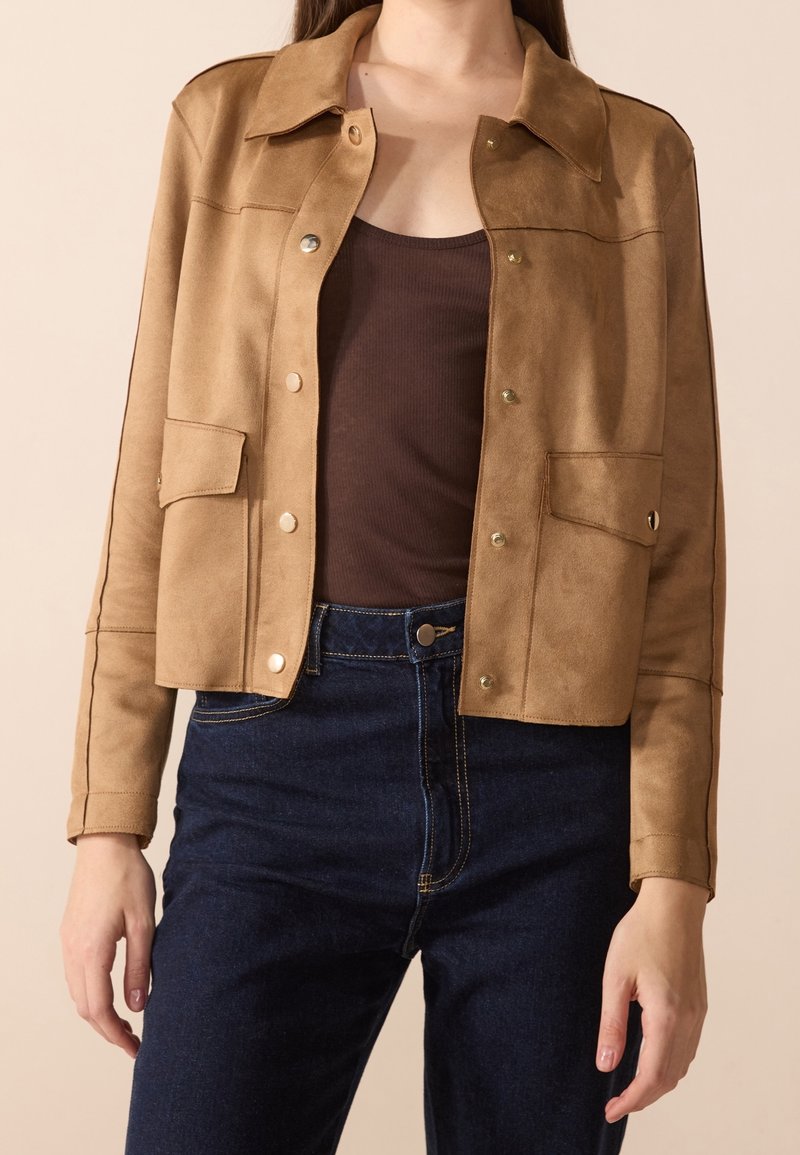 Veste en daim marron avec un col, des boutons-pression et deux poches avant. Associée à un jeans taille haute bleu foncé. Texture lisse.