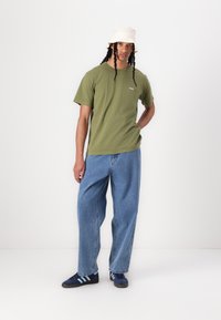 Olive green cotton t-shirt, loose-fit blue denim jeans, and navy sneakers with light blue stripes. Beige bucket hat completes the look.