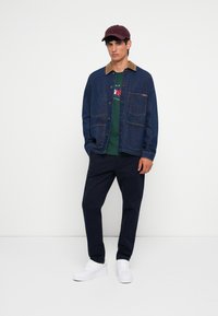 Chaqueta de mezclilla con un collar marrón, camiseta gráfica verde debajo, pantalones azul marino y zapatillas blancas. Una gorra burdeos completa el atuendo.