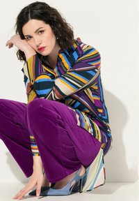 Multicolore gestreepte overhemd met knopen in blauw, geel en paars, gecombineerd met paarse ribfluwelen broeken en lichtblauwe hakken.