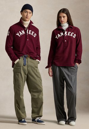 Hnědé košile na knoflíky s textem "YANKEES", olivové cargo kalhoty, šedé kalhoty širokého střihu, námořnické a bílé boty, béžové texturované pozadí.