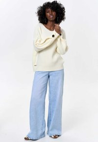 Cream-colored, ribbed knit sweater with oversized sleeves and black buttons, paired with light blue wide-leg jeans. Textured and casual.
