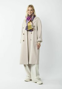 Beige wollen blend lange jas met dubbele knopen, gelaagd over een lichtbeige top en brede crème broek. Kleurrijke geruite sjaal.