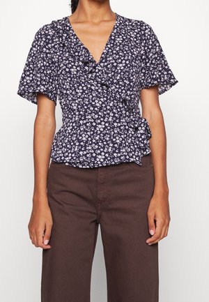 Femme portant un chemisier cache-cœur bleu marine à imprimé floral blanc et manches volantées, associé à un pantalon taille haute marron foncé.