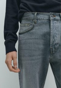 Jeansy dżinsowe w jasnoszarym kolorze z lekkim wyblaknięciem, z pięcioma kieszeniami i akcentami przeszyć. Noszone z ciemnym swetrem.