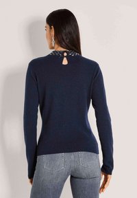 Frau mit glattem dunkelbraunem Haar, die einen marineblauen Pullover mit einem kleinen Schlüssel-Ausschnitt und gestrickten Details am hinteren Halsausschnitt trägt, kombiniert mit grauen Jeans.