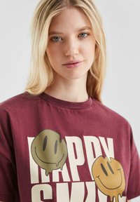 Blonde vrouw in een kastanjebruine T-shirt met grote witte tekst en twee druipende smiley-gezichten in groen en geel.