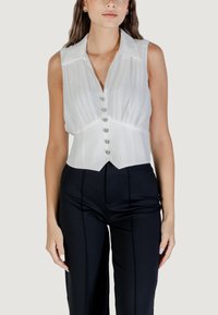 Blusa bianca senza maniche con colletto strutturato, design plissettato e bottoni argentati sul davanti, abbinata a pantaloni neri a vita alta.
