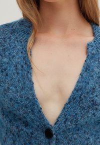 Zbliżenie osoby noszącej niebieski kardigan o teksturowanym splocie, z zapiętym pojedynczym czarnym guzikiem, ukazujące linię dekoltu i obojczyk.