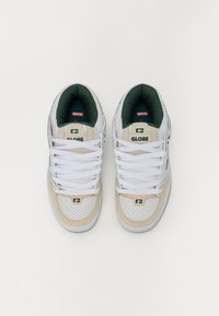 White and beige sneakers with a perforated toe, green inner lining, and flat white laces. Features a logo on the tongue and side.