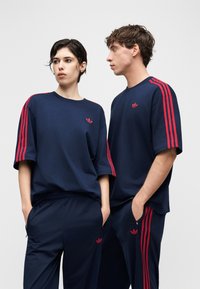 Due persone indossano tute Adidas blu navy abbinate con strisce rosse e loghi, in piedi con le mani nelle tasche contro uno sfondo semplice.
