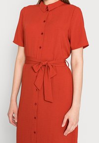 Abito rosso ruggine a maniche corte con chiusura a bottoni, cintura in vita e colletto. Realizzato in un tessuto texturizzato con una finitura liscia.