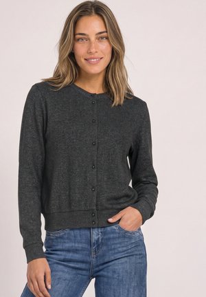 Femme souriante aux cheveux mi-longs porte un cardigan boutonné gris foncé et un jean bleu avec une main dans la poche.