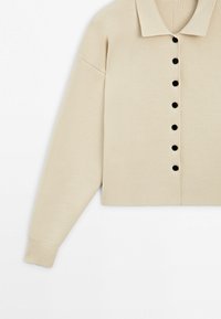 Beige ribbed knit cardigan with long sleeves, black buttons down the front, and a wide collar.