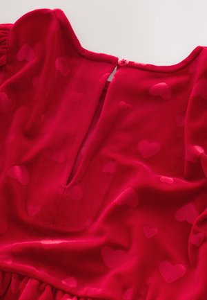 Rode fluwelen jurk met een hartjespatroon, ronde halslijn, knoopsluiting aan de achterkant en een zachte textuur. De geplooide taille accentueert de silhouet.