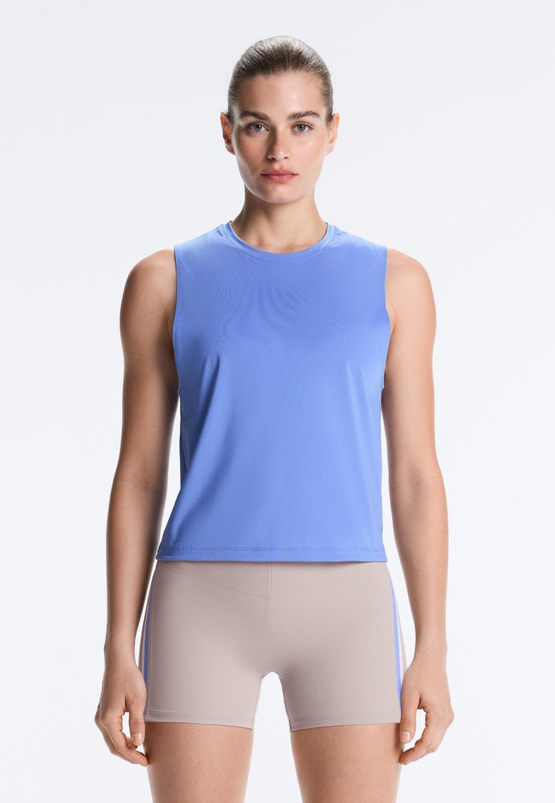 Femme portant un débardeur bleu et un short beige ajusté, debout droite sur un fond blanc uni.