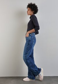 Mörk långärmad topp, ljusblå vida jeans med fem fickor och vita sneakers med rosa detaljer. Sido vy mot en neutral bakgrund.