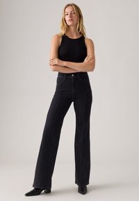 Mujer de pie con los brazos cruzados, vistiendo una blusa sin mangas negra, jeans negros de cintura alta y botines negros de punta.