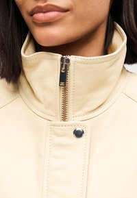 Κρεμ δερμάτινο μπουφάν με ψηλό γιακά, ασημένιο φερμουάρ και λεπτομέρεια με κουμπί κούμπωμα. Ομαλή υφή με διακοσμητικές ραφές.