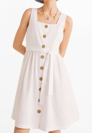 Mouwloze witte jurk met vierkante halslijn, houten knopen aan de voorkant, tailleceintuur, gedragen met een kleurrijke kralenhalsketting.