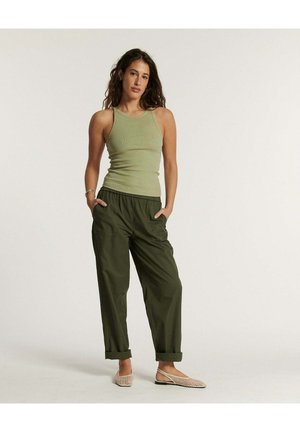 Femme debout, mains dans les poches, portant un haut sans manches vert olive, un pantalon ample vert foncé, et des chaussures plates en tissu beige sur fond blanc.