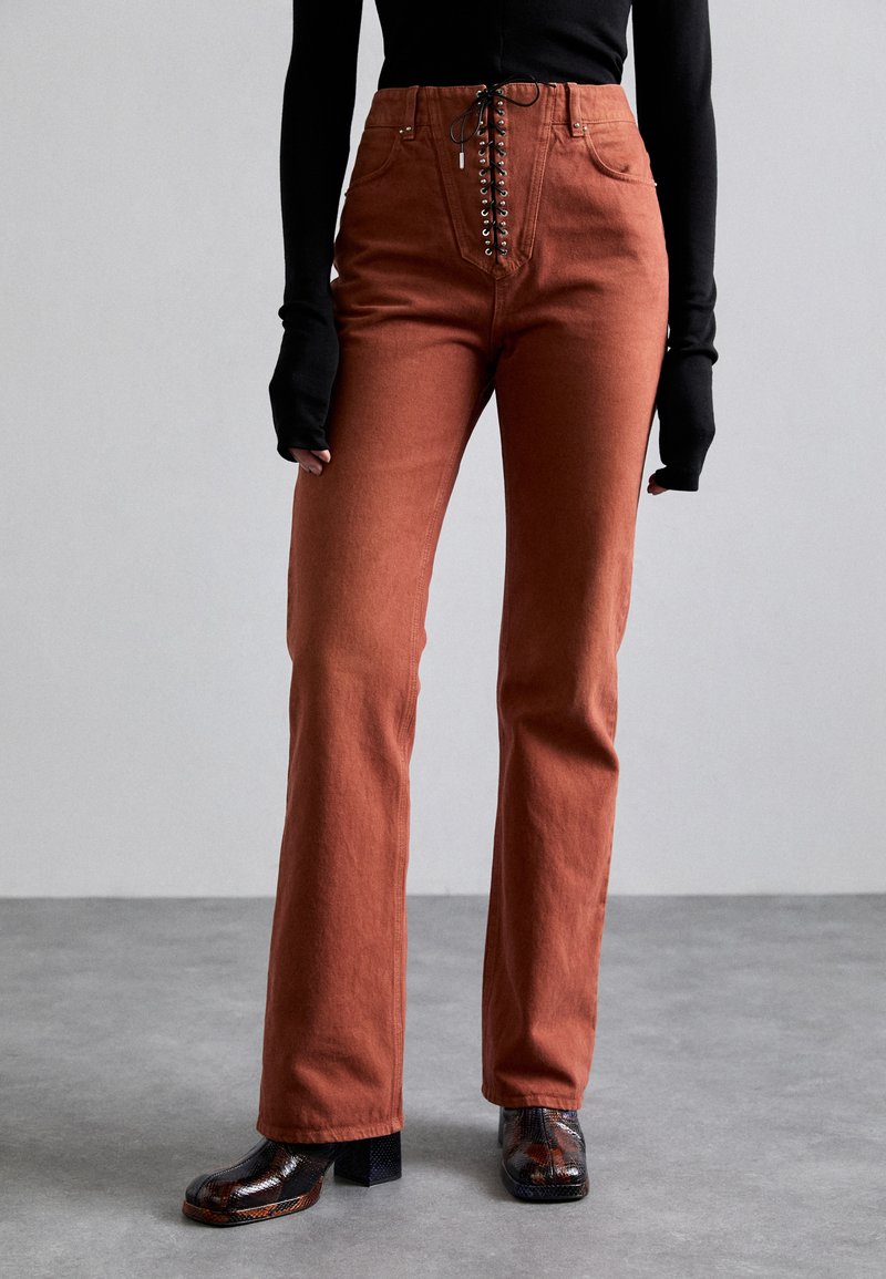 Russetkleurige hooggezette broek met vetersluiting aan de voorkant, gestructureerde vorm en zachte textuur, in combinatie met gestreepte blokhak schoenen.