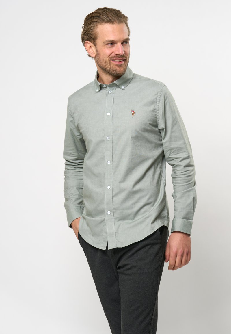 Camicia verde chiaro con colletto, dettaglio ricamato sul petto e maniche lunghe. Indossata con pantaloni scuri. Tessuto dalla superficie liscia.