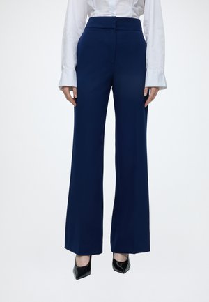 Femme portant un pantalon large taille haute bleu marine, une chemise blanche à manches longues avec des poignets évasés, et des talons noirs à bouts pointus.