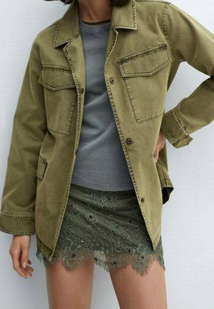 Personne portant une veste vert olive par-dessus une chemise grise et une jupe en dentelle vert foncé avec broderie florale et détails en perles.