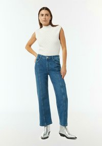 Bezrekawnik dzianinowy w kolorze kremowym z prążkowaną teksturą, zestawiony z niebieskimi spodniami dżinsowymi z wysokim stanem i prostą nogawką. Srebrne botki za kostkę.