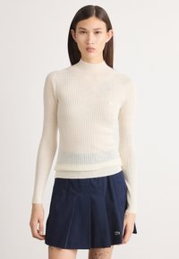 Suéter de malha canelada em creme, com gola alta e mangas longas, apresentando um design justo, combinado com uma saia plissada azul-marinho. Detalhe de logótipo notável.