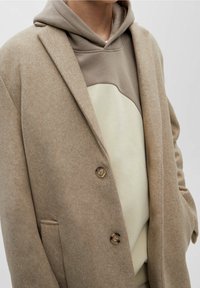 Manteau en laine beige à revers crantés, deux boutons et poches latérales, porté sur un sweat à capuche pastel avec un motif abstrait crème.