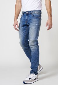 Vaqueros de denim azul desgastado con detalles rasgados, corte ajustado, diseño de cinco bolsillos y cintura estándar, combinados con zapatillas blancas y grises.