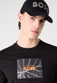 Melna T-kreka ar baltu lapu grafiku un oranžiem burtiem, kas lasa "THROUGH THE JUNGLE BOSS". Melna cepure ar iegravētu "BOSS" logotipu.
