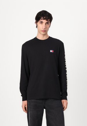 Black long-sleeve t-shirt made of cotton. Features a small logo patch on the chest and text in a light color along the sleeve.