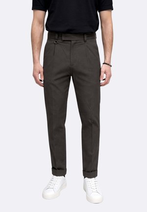 Uomo indossa pantaloni slim fit grigio scuro con orli risvoltati, passanti per cintura neri e sneakers bianche, in piedi davanti a uno sfondo semplice.