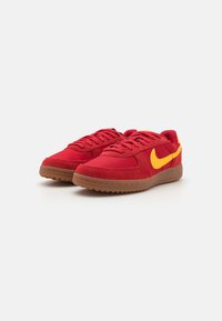 Rote Wildleder- und Mesh-Sneaker mit einem gelben Nike-Swoosh, ausgestattet mit einer braunen Gummisohle und einem abgerundeten Zehendesign. Mit Schnürverschluss und gepolstertem Kragen.