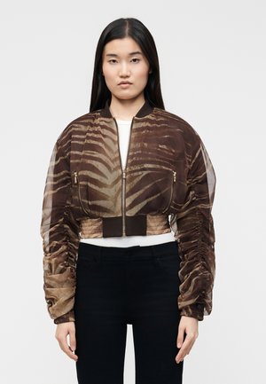 Donna con capelli lunghi neri che indossa una giacca a rete marrone e beige con stampa zebrita e maniche arricciate sopra una maglia bianca e pantaloni neri.