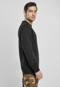 Melnā džemperis no mīkstas auduma, ar apaļu kakla izgriezumu un garām piedurknēm, kopā ar haki kamuflāžas biksēm.